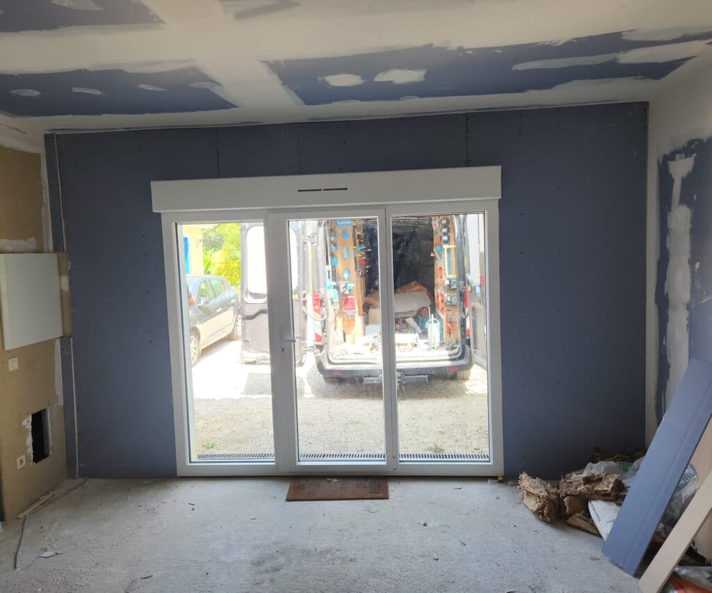 Pièce en rénovation avec une baie vitrée blanche à double vantail donnant sur l'extérieur, vue sur un fourgon d'artisan ouvert et du matériel de chantier.