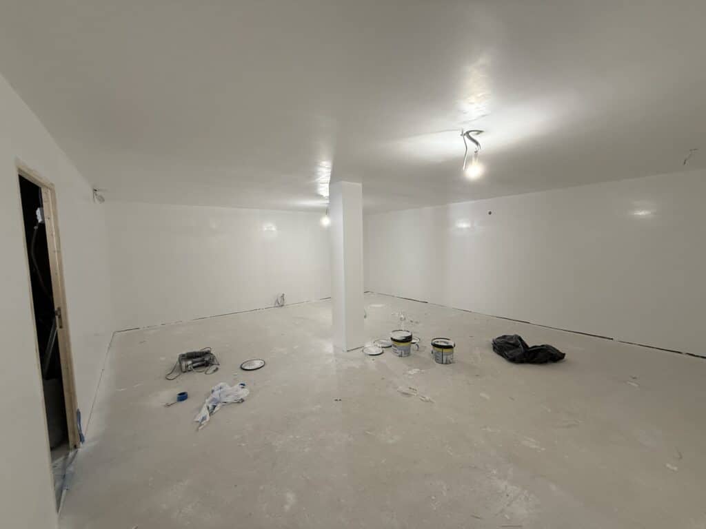 Intérieur d'une pièce vide en cours de rénovation, murs et plafond blancs fraîchement peints, sol brut avec pots de peinture et outils.