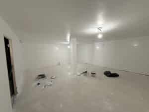 Intérieur d'une pièce vide en cours de rénovation, murs et plafond blancs fraîchement peints, sol brut avec pots de peinture et outils.