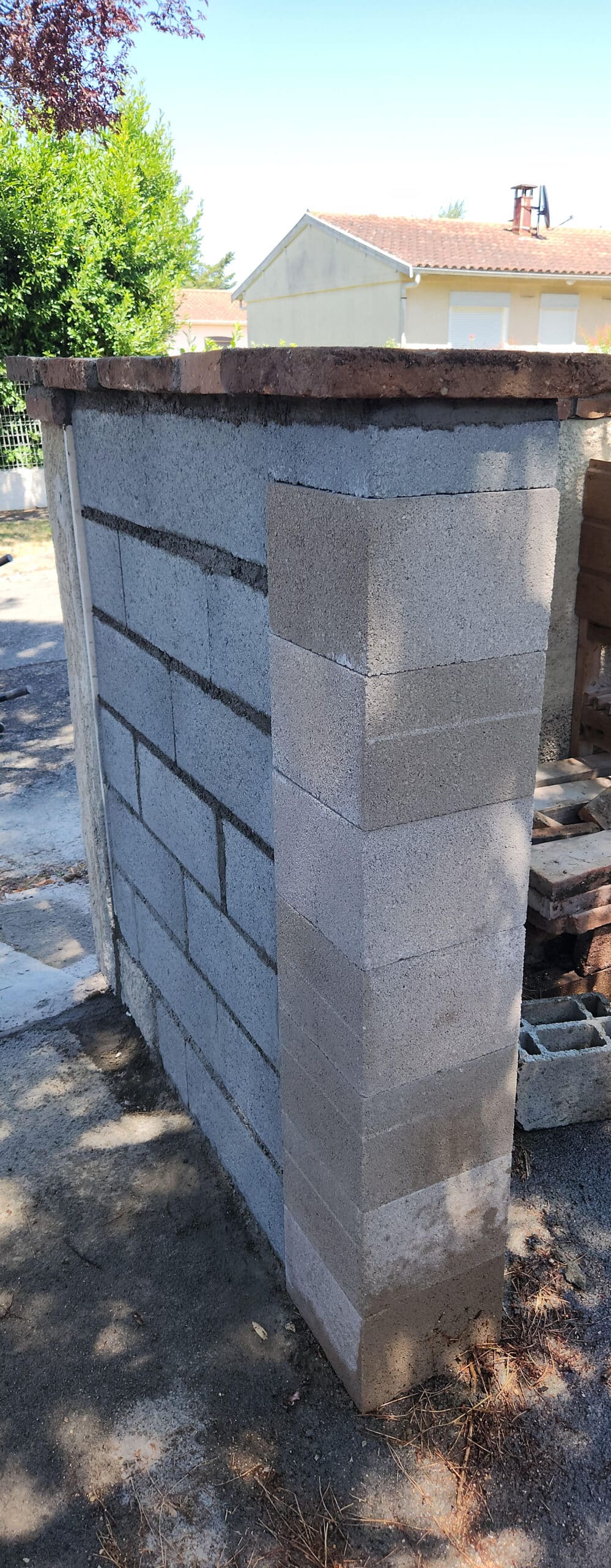 Mur en parpaings en cours de construction avec poteau d'angle et chapeau de finition en brique sous le soleil.